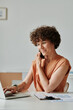 © AnnaStills - Young businesswoman with curly hair talking on mobile phone and working on laptop at her workplace at office