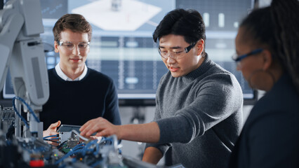 Male Student Holding a Tablet And Pointing at Robotic Hand While Working on Project with Asian Classmate and Black Teacher. Modern Technologies, Science and Developing at University Concept.