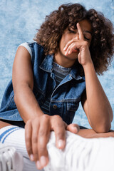 Wall Mural - brunette african american woman in denim vest obscuring face with hand on blue background