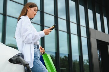 Happy young woman standing on city parking near electric car, charging automobile battery from small city station, holding shopping bags.