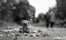 Lost Teddy Bear With Sad Face Sitting On Footpath With Blurry People Lonely Bear Doll Sit Down On The Brick Floor In Gloomy Day, Lost Toy Or Loneliness Concept, International Missing Children