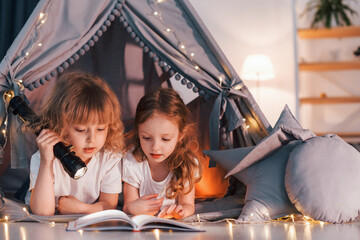 Wall Mural - Reading book and using flashlight. Two little girls is in the tent in domestic room together