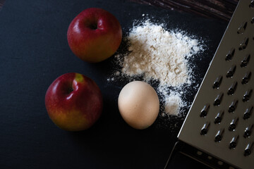 Ingredients for the recipe for apple pancakes, muffins on black graphite table: two red apples, egg, wheat flour and a grater