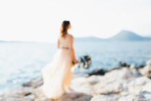 Silhouette Of Lovely Bride In A Pastel Wedding Dress Stands On A Rock Above The Sea With A Bouquet Of Flowers In Her Hands