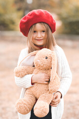  Smiling funny kid girl 4-5 year old playing with fluffy rabbit toy outdoors. Child toddler wear red beret and knit sweater in park. Cheerful little pupil. Childhood. Autumn season.