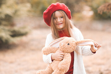 Smiling funny kid girl 4-5 year old playing with fluffy rabbit toy outdoors. Child toddler wear red beret and knit sweater in park. Cheerful little pupil. Childhood. Autumn season.