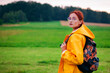© Тамара Печеная - Portrait of a female tourist with a backpack in glasses wearing yellow coat. Young woman travels during summer or autumn season. Hitch-hiking, trip and travelling concept.