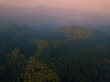 A Panoramic View Of The Foggy Mountains With Dense Of Forest. Orange Sky With Clouds Over Layers Of Green Hills And Mountains. Copy Space.  Menoreh Hill, Central Java, Indonesia