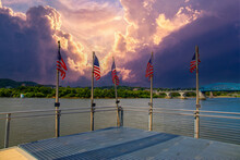 American Flags Flying On The Edge Of The Pier Surrounded By The Flowing Waters Of The Tennessee River And Lush Green Trees With Blue Sky And Powerful Clouds At Sunset At Ross's Landing In Chattanooga