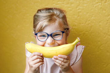 Happy little child girl with yellow banana like smile on yellow background. Preschool girl with glasses smiling. Healthy fruits for children