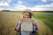 © olga_sova - A beautiful middle-aged farmer woman in a straw hat and a plaid shirt stands in a field of golden ripening wheat during the daytime in the sunlight