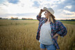 © olga_sova - A beautiful middle-aged farmer woman in a straw hat and a plaid shirt stands in a field of golden ripening wheat during the daytime in the sunlight