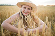 © olga_sova - A beautiful middle-aged farmer woman in a straw hat and a plaid shirt stands in a field of golden ripening wheat during the daytime in the sunlight