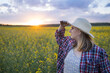 © olga_sova - A beautiful middle-aged farmer woman in a straw hat and a plaid shirt stands in a field of flowering rapeseed in the sunlight