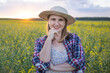 © olga_sova - A beautiful middle-aged farmer woman in a straw hat and a plaid shirt stands in a field of flowering rapeseed in the sunlight