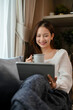 © bongkarn - Beautiful young Asian female sipping coffee while watching movies on tablet in the living room.