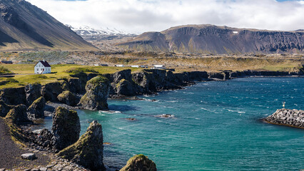 Leinwandbilder - Amazing nature of Iceland. View of the basalt rocks formation on the coastline with beautiful houses and the Mountain Stapafell in the background at Arnarstapi Village in summer sunny day in Iceland
