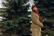 Positive woman wearing warm coat and hat with scarf dancing and having fun in winter park with coniferous fir trees while looking at camera 