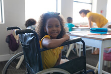 Portrait Of Smiling African American Elementary Girl Sitting On Wheelchair At Desk In Classroom