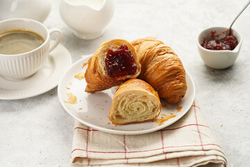 Two french soft croissants with jam on a white plate on a checkered napkin, a cup of black coffee in white sophisticated cup, milk, sugar