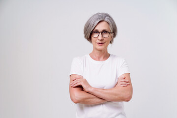 Beautiful mature grey haired woman, serious businesswoman with folded hands looking straight in camera wearing white t-shirt isolated on white background. Mature people beauty concept.