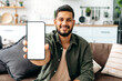 © Kateryna - Mock-up and copy-space concept. Positive indian or arabian guy in casual wear, sits on sofa in living room, shows smart phone with empty white mock-up screen, for advertisement or presentation, smiles