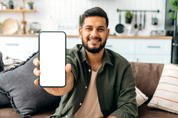 Mock-up and copy-space concept. Positive indian or arabian guy in casual wear, sits on sofa in living room, shows smart phone with empty white mock-up screen, for advertisement or presentation, smiles