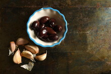 Dark Olives In Small Bowl With Blue Trim Beside Garlic Cloves On Dark Grunge Background.