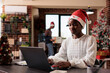 © DC Studio - African american woman with santa hat working on business in office with christmas tree and lights. Festive employee using laptop at company job with seasonal decor and ornaments.