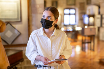 Portrait of young female visitor in protective face mask with paper guide at paintings exposition