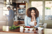 Portrait Of A Happy, Healthy And Carefree Young Woman Preparing A Healthy Meal At Home With Her Husband In The Background. Black Wife Making An Organic Vegetarian Salad For Lunch In A Kitchen
