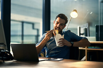 Canvas Print - Young hungry businessman working late and eating at desk. Man having takeout food in the office at work station in the evening. Male entrepreneur eating asian meal during the night at the workplace