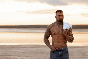 Handsome young muscular italian man posing shirtless on the beach, sunset summer time