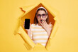 © sementsova321 - Indoor shot of shocked surprised woman wearing striped t shirt looking through breakthrough of yellow background, showing blank screen of smart phone, covering mouth with palm.