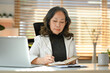 © Prathankarnpap - Concentrated middle aged businesswoman sitting in front of laptop and writing important information on notebook