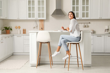 Beautiful young woman with notebook sitting on stool in kitchen