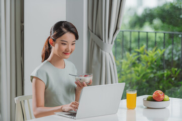 Wall Mural - Portrait of beautiful young woman working with laptop while eating at home.