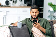 © Kateryna - Calm indian or arabian attractive guy using his smartphone while sitting on sofa in living room, browsing internet and social networks, texting with friends or family, holds a cup of coffee in hand
