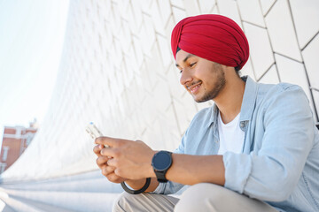 portrait of cheerful young indian guy in turban sending message on mobile phone sitting on the steps