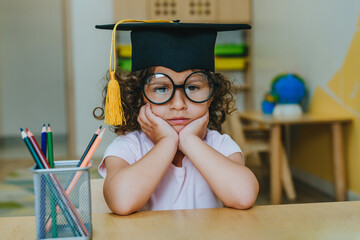 Portrait of unhappy little girl in graduation mortar hat