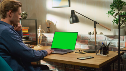 Fototapete - Young Handsome Man Working from Home on Laptop Computer with Green Screen Mock Up Display. Creative Male Checking Social Media, Browsing Internet. Living Room in Bright Loft Apartment.