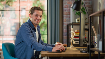 Poster - Portrait of a Young Handsome Man Sitting at Home, Working on Desktop Computer. Creative Male Looking Directly at Camera and Smiles. Living Room with Big Window in Loft Apartment.