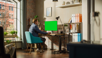 Fototapete - Young Handsome Man Working from Home on Desktop Computer with Green Screen Mock Up Display. Creative Male Checking Social Media, Browsing Internet. Living Room in Bright Loft Apartment.