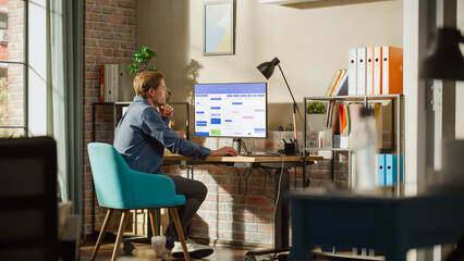 Fototapete - Remote Work Concept: Man Using Desktop Computer With Office Software to Check Schedule in Calendar. Young Male Planning Business Meetings in Stylish Loft Apartment with Big Window.