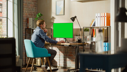 Leinwandbilder - Young Handsome Man Working from Home on Desktop Computer with Green Screen Mock Up Display. Male Checking Corporate Accounts, Messaging Colleagues. Loft Living Room with Big Window.