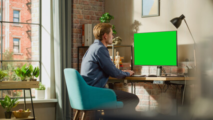 Poster - Young Handsome Man Working from Home on Desktop Computer with Green Screen Mock Up Display. Male Checking Corporate Accounts, Messaging Colleagues. Loft Living Room with Big Window.