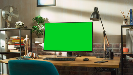 Fototapete - Desktop Computer Monitor Standing on a Desk with a Green Screen Chromakey Mock Up Display. Cozy Empty Loft Apartment with a Lamp, Notebooks, Smartphone and Headphones on the Table.