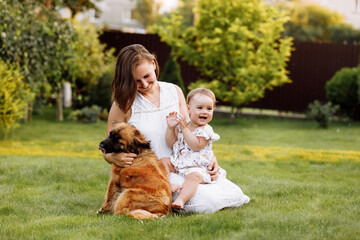 Family day, mother's day. Beautiful smiling young mom and baby daughter cuddling happy domestic dog on the backyard lawn.Mother with child girl are having fun with pet outdoors on summer holiday.