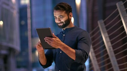 Smiling young indian business man professional, eastern businessman executive standing outdoors on street reading ebook holding using digital tablet online technology in night city with urbans lights.