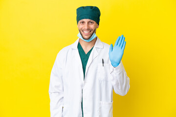 Wall Mural - Surgeon blonde man in green uniform isolated on yellow background saluting with hand with happy expression